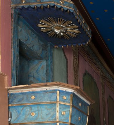 Zdjęcie nr 1: Ambona wisząca, przyścienna, z wejściem od strony zakrystii. Kosz jest na planie czworoboku o ściętych narożach, ścianki są o wklęsło-wypukłej powierzchni. Podwieszenie jest w formie profilowanego, zwężającego się ku dołowi gzymsu. W zaplecku ambony znajduje się prostokątny otwór wejściowy ujęty parą pilastrów o kanelowanych trzonach. Płytki baldachim, na rzucie czworoboku o ściętych narożach, nakryty jest spłaszczonym hełmem. Kosz ambony zdobią pojedyncze rozetki, krawędzie ujmują wąskie, żłobkowane listwy. Wzdłuż krawędzi baldachimu znajdują się pojedyncze rozetki oraz motyw lambrekinu. Podniebie zdobi płaskorzeźbiona gołębica Ducha Świętego ujęta srebrzonym wieńcem obłoków i kolistą, promienistą glorią. Struktura ambony jest marmoryzowana w odcieniach błękitu i szarości, ornamentyka jest złocona i srebrzona.
