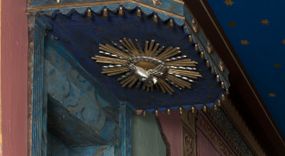 Zdjęcie nr 1: Ambona wisząca, przyścienna, z wejściem od strony zakrystii. Kosz jest na planie czworoboku o ściętych narożach, ścianki są o wklęsło-wypukłej powierzchni. Podwieszenie jest w formie profilowanego, zwężającego się ku dołowi gzymsu. W zaplecku ambony znajduje się prostokątny otwór wejściowy ujęty parą pilastrów o kanelowanych trzonach. Płytki baldachim, na rzucie czworoboku o ściętych narożach, nakryty jest spłaszczonym hełmem. Kosz ambony zdobią pojedyncze rozetki, krawędzie ujmują wąskie, żłobkowane listwy. Wzdłuż krawędzi baldachimu znajdują się pojedyncze rozetki oraz motyw lambrekinu. Podniebie zdobi płaskorzeźbiona gołębica Ducha Świętego ujęta srebrzonym wieńcem obłoków i kolistą, promienistą glorią. Struktura ambony jest marmoryzowana w odcieniach błękitu i szarości, ornamentyka jest złocona i srebrzona.
