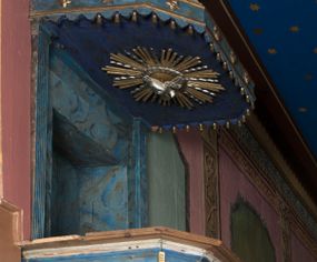 Zdjęcie nr 1: Ambona wisząca, przyścienna, z wejściem od strony zakrystii. Kosz jest na planie czworoboku o ściętych narożach, ścianki są o wklęsło-wypukłej powierzchni. Podwieszenie jest w formie profilowanego, zwężającego się ku dołowi gzymsu. W zaplecku ambony znajduje się prostokątny otwór wejściowy ujęty parą pilastrów o kanelowanych trzonach. Płytki baldachim, na rzucie czworoboku o ściętych narożach, nakryty jest spłaszczonym hełmem. Kosz ambony zdobią pojedyncze rozetki, krawędzie ujmują wąskie, żłobkowane listwy. Wzdłuż krawędzi baldachimu znajdują się pojedyncze rozetki oraz motyw lambrekinu. Podniebie zdobi płaskorzeźbiona gołębica Ducha Świętego ujęta srebrzonym wieńcem obłoków i kolistą, promienistą glorią. Struktura ambony jest marmoryzowana w odcieniach błękitu i szarości, ornamentyka jest złocona i srebrzona.
