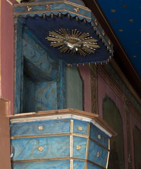 Zdjęcie nr 1: Ambona wisząca, przyścienna, z wejściem od strony zakrystii. Kosz jest na planie czworoboku o ściętych narożach, ścianki są o wklęsło-wypukłej powierzchni. Podwieszenie jest w formie profilowanego, zwężającego się ku dołowi gzymsu. W zaplecku ambony znajduje się prostokątny otwór wejściowy ujęty parą pilastrów o kanelowanych trzonach. Płytki baldachim, na rzucie czworoboku o ściętych narożach, nakryty jest spłaszczonym hełmem. Kosz ambony zdobią pojedyncze rozetki, krawędzie ujmują wąskie, żłobkowane listwy. Wzdłuż krawędzi baldachimu znajdują się pojedyncze rozetki oraz motyw lambrekinu. Podniebie zdobi płaskorzeźbiona gołębica Ducha Świętego ujęta srebrzonym wieńcem obłoków i kolistą, promienistą glorią. Struktura ambony jest marmoryzowana w odcieniach błękitu i szarości, ornamentyka jest złocona i srebrzona.
