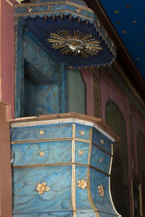 Zdjęcie nr 1: Ambona wisząca, przyścienna, z wejściem od strony zakrystii. Kosz jest na planie czworoboku o ściętych narożach, ścianki są o wklęsło-wypukłej powierzchni. Podwieszenie jest w formie profilowanego, zwężającego się ku dołowi gzymsu. W zaplecku ambony znajduje się prostokątny otwór wejściowy ujęty parą pilastrów o kanelowanych trzonach. Płytki baldachim, na rzucie czworoboku o ściętych narożach, nakryty jest spłaszczonym hełmem. Kosz ambony zdobią pojedyncze rozetki, krawędzie ujmują wąskie, żłobkowane listwy. Wzdłuż krawędzi baldachimu znajdują się pojedyncze rozetki oraz motyw lambrekinu. Podniebie zdobi płaskorzeźbiona gołębica Ducha Świętego ujęta srebrzonym wieńcem obłoków i kolistą, promienistą glorią. Struktura ambony jest marmoryzowana w odcieniach błękitu i szarości, ornamentyka jest złocona i srebrzona.
