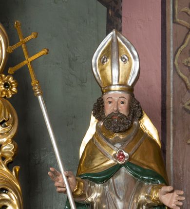 Zdjęcie nr 6: Figura przedstawiająca św. Wojciecha polichromowana, niemal pełnoplastyczna, wydrążona z tyłu. Święty ukazany w pozycji stojącej, w hieratycznej pozie. Głowa zwrócona lekko w prawą stronę, ręce wysunięte przed siebie, w prawej dłoni krzyż patriarchalny. Twarz szeroka, schematyczne opracowana, okolona ciemnobrązowymi włosami i zarostem. Święty jest ubrany w szaty biskupie: czerwoną sutannę, srebrną komżę oraz złotą kapę z zielonym podszyciem spiętą na piersi broszą, na głowie infuła.