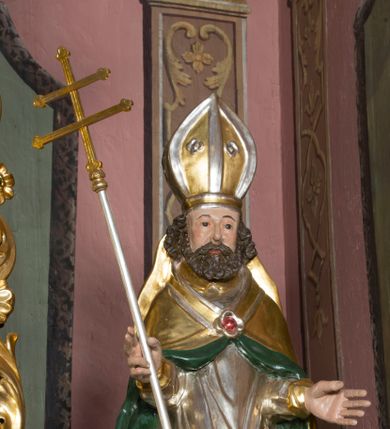 Zdjęcie nr 5: Figura przedstawiająca św. Wojciecha polichromowana, niemal pełnoplastyczna, wydrążona z tyłu. Święty ukazany w pozycji stojącej, w hieratycznej pozie. Głowa zwrócona lekko w prawą stronę, ręce wysunięte przed siebie, w prawej dłoni krzyż patriarchalny. Twarz szeroka, schematyczne opracowana, okolona ciemnobrązowymi włosami i zarostem. Święty jest ubrany w szaty biskupie: czerwoną sutannę, srebrną komżę oraz złotą kapę z zielonym podszyciem spiętą na piersi broszą, na głowie infuła.