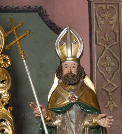 Zdjęcie nr 4: Figura przedstawiająca św. Wojciecha polichromowana, niemal pełnoplastyczna, wydrążona z tyłu. Święty ukazany w pozycji stojącej, w hieratycznej pozie. Głowa zwrócona lekko w prawą stronę, ręce wysunięte przed siebie, w prawej dłoni krzyż patriarchalny. Twarz szeroka, schematyczne opracowana, okolona ciemnobrązowymi włosami i zarostem. Święty jest ubrany w szaty biskupie: czerwoną sutannę, srebrną komżę oraz złotą kapę z zielonym podszyciem spiętą na piersi broszą, na głowie infuła.