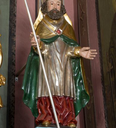 Zdjęcie nr 3: Figura przedstawiająca św. Wojciecha polichromowana, niemal pełnoplastyczna, wydrążona z tyłu. Święty ukazany w pozycji stojącej, w hieratycznej pozie. Głowa zwrócona lekko w prawą stronę, ręce wysunięte przed siebie, w prawej dłoni krzyż patriarchalny. Twarz szeroka, schematyczne opracowana, okolona ciemnobrązowymi włosami i zarostem. Święty jest ubrany w szaty biskupie: czerwoną sutannę, srebrną komżę oraz złotą kapę z zielonym podszyciem spiętą na piersi broszą, na głowie infuła.