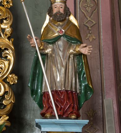 Zdjęcie nr 2: Figura przedstawiająca św. Wojciecha polichromowana, niemal pełnoplastyczna, wydrążona z tyłu. Święty ukazany w pozycji stojącej, w hieratycznej pozie. Głowa zwrócona lekko w prawą stronę, ręce wysunięte przed siebie, w prawej dłoni krzyż patriarchalny. Twarz szeroka, schematyczne opracowana, okolona ciemnobrązowymi włosami i zarostem. Święty jest ubrany w szaty biskupie: czerwoną sutannę, srebrną komżę oraz złotą kapę z zielonym podszyciem spiętą na piersi broszą, na głowie infuła.