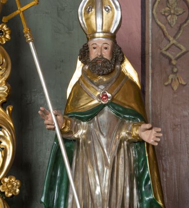 Zdjęcie nr 1: Figura przedstawiająca św. Wojciecha polichromowana, niemal pełnoplastyczna, wydrążona z tyłu. Święty ukazany w pozycji stojącej, w hieratycznej pozie. Głowa zwrócona lekko w prawą stronę, ręce wysunięte przed siebie, w prawej dłoni krzyż patriarchalny. Twarz szeroka, schematyczne opracowana, okolona ciemnobrązowymi włosami i zarostem. Święty jest ubrany w szaty biskupie: czerwoną sutannę, srebrną komżę oraz złotą kapę z zielonym podszyciem spiętą na piersi broszą, na głowie infuła.
