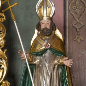 Zdjęcie nr 1: Figura przedstawiająca św. Wojciecha polichromowana, niemal pełnoplastyczna, wydrążona z tyłu. Święty ukazany w pozycji stojącej, w hieratycznej pozie. Głowa zwrócona lekko w prawą stronę, ręce wysunięte przed siebie, w prawej dłoni krzyż patriarchalny. Twarz szeroka, schematyczne opracowana, okolona ciemnobrązowymi włosami i zarostem. Święty jest ubrany w szaty biskupie: czerwoną sutannę, srebrną komżę oraz złotą kapę z zielonym podszyciem spiętą na piersi broszą, na głowie infuła.