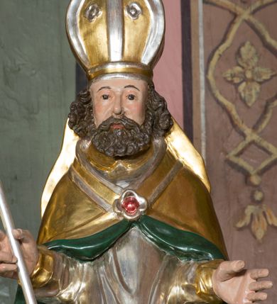 Zdjęcie nr 8: Figura przedstawiająca św. Wojciecha polichromowana, niemal pełnoplastyczna, wydrążona z tyłu. Święty ukazany w pozycji stojącej, w hieratycznej pozie. Głowa zwrócona lekko w prawą stronę, ręce wysunięte przed siebie, w prawej dłoni krzyż patriarchalny. Twarz szeroka, schematyczne opracowana, okolona ciemnobrązowymi włosami i zarostem. Święty jest ubrany w szaty biskupie: czerwoną sutannę, srebrną komżę oraz złotą kapę z zielonym podszyciem spiętą na piersi broszą, na głowie infuła.