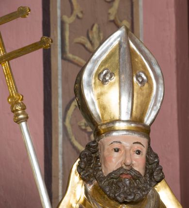 Zdjęcie nr 10: Figura przedstawiająca św. Wojciecha polichromowana, niemal pełnoplastyczna, wydrążona z tyłu. Święty ukazany w pozycji stojącej, w hieratycznej pozie. Głowa zwrócona lekko w prawą stronę, ręce wysunięte przed siebie, w prawej dłoni krzyż patriarchalny. Twarz szeroka, schematyczne opracowana, okolona ciemnobrązowymi włosami i zarostem. Święty jest ubrany w szaty biskupie: czerwoną sutannę, srebrną komżę oraz złotą kapę z zielonym podszyciem spiętą na piersi broszą, na głowie infuła.