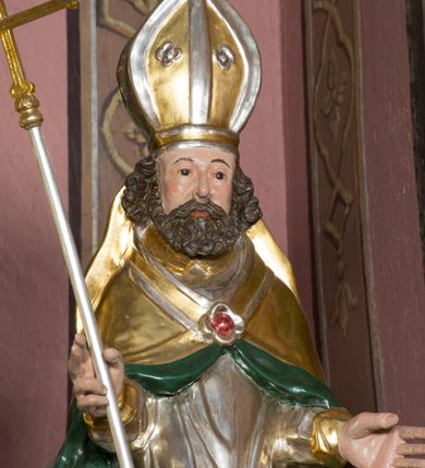 Zdjęcie nr 7: Figura przedstawiająca św. Wojciecha polichromowana, niemal pełnoplastyczna, wydrążona z tyłu. Święty ukazany w pozycji stojącej, w hieratycznej pozie. Głowa zwrócona lekko w prawą stronę, ręce wysunięte przed siebie, w prawej dłoni krzyż patriarchalny. Twarz szeroka, schematyczne opracowana, okolona ciemnobrązowymi włosami i zarostem. Święty jest ubrany w szaty biskupie: czerwoną sutannę, srebrną komżę oraz złotą kapę z zielonym podszyciem spiętą na piersi broszą, na głowie infuła.