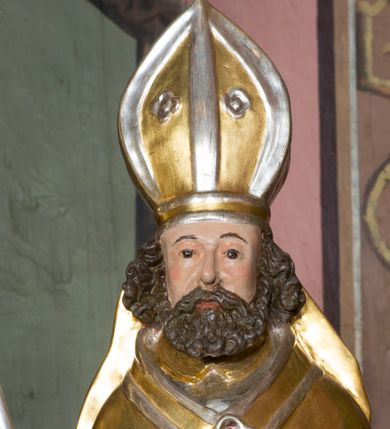 Zdjęcie nr 9: Figura przedstawiająca św. Wojciecha polichromowana, niemal pełnoplastyczna, wydrążona z tyłu. Święty ukazany w pozycji stojącej, w hieratycznej pozie. Głowa zwrócona lekko w prawą stronę, ręce wysunięte przed siebie, w prawej dłoni krzyż patriarchalny. Twarz szeroka, schematyczne opracowana, okolona ciemnobrązowymi włosami i zarostem. Święty jest ubrany w szaty biskupie: czerwoną sutannę, srebrną komżę oraz złotą kapę z zielonym podszyciem spiętą na piersi broszą, na głowie infuła.