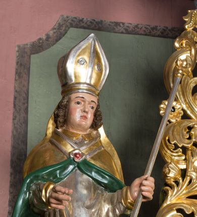 Zdjęcie nr 4: Figura przedstawiająca św. Stanisława polichromowana, niemal pełnoplastyczna, wydrążona z tyłu. Święty ukazany w pozycji stojącej, w hieratycznej pozie. Głowa przechylona lekko w lewą stronę, ręce wysunięte, skierowane w lewą stronę, w lewej dłoni święty trzyma pastorał. Twarz szeroka, schematycznie opracowana, z puklami ciemnobrązowych włosów po bokach. Święty jest ubrany w szaty biskupie: czerwoną sutannę, srebrną komżę oraz złotą kapę z zielonym podszyciem spiętą na piersi broszą, na głowie infuła.