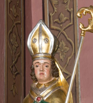 Zdjęcie nr 5: Figura przedstawiająca św. Stanisława polichromowana, niemal pełnoplastyczna, wydrążona z tyłu. Święty ukazany w pozycji stojącej, w hieratycznej pozie. Głowa przechylona lekko w lewą stronę, ręce wysunięte, skierowane w lewą stronę, w lewej dłoni święty trzyma pastorał. Twarz szeroka, schematycznie opracowana, z puklami ciemnobrązowych włosów po bokach. Święty jest ubrany w szaty biskupie: czerwoną sutannę, srebrną komżę oraz złotą kapę z zielonym podszyciem spiętą na piersi broszą, na głowie infuła.