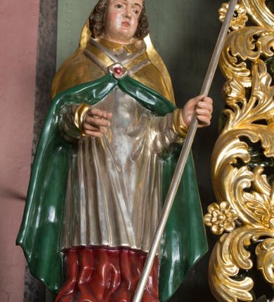 Zdjęcie nr 2: Figura przedstawiająca św. Stanisława polichromowana, niemal pełnoplastyczna, wydrążona z tyłu. Święty ukazany w pozycji stojącej, w hieratycznej pozie. Głowa przechylona lekko w lewą stronę, ręce wysunięte, skierowane w lewą stronę, w lewej dłoni święty trzyma pastorał. Twarz szeroka, schematycznie opracowana, z puklami ciemnobrązowych włosów po bokach. Święty jest ubrany w szaty biskupie: czerwoną sutannę, srebrną komżę oraz złotą kapę z zielonym podszyciem spiętą na piersi broszą, na głowie infuła.