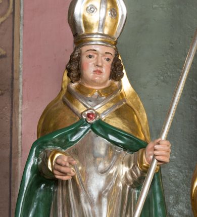 Zdjęcie nr 1: Figura przedstawiająca św. Stanisława polichromowana, niemal pełnoplastyczna, wydrążona z tyłu. Święty ukazany w pozycji stojącej, w hieratycznej pozie. Głowa przechylona lekko w lewą stronę, ręce wysunięte, skierowane w lewą stronę, w lewej dłoni święty trzyma pastorał. Twarz szeroka, schematycznie opracowana, z puklami ciemnobrązowych włosów po bokach. Święty jest ubrany w szaty biskupie: czerwoną sutannę, srebrną komżę oraz złotą kapę z zielonym podszyciem spiętą na piersi broszą, na głowie infuła.
