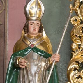 Zdjęcie nr 1: Figura przedstawiająca św. Stanisława polichromowana, niemal pełnoplastyczna, wydrążona z tyłu. Święty ukazany w pozycji stojącej, w hieratycznej pozie. Głowa przechylona lekko w lewą stronę, ręce wysunięte, skierowane w lewą stronę, w lewej dłoni święty trzyma pastorał. Twarz szeroka, schematycznie opracowana, z puklami ciemnobrązowych włosów po bokach. Święty jest ubrany w szaty biskupie: czerwoną sutannę, srebrną komżę oraz złotą kapę z zielonym podszyciem spiętą na piersi broszą, na głowie infuła.