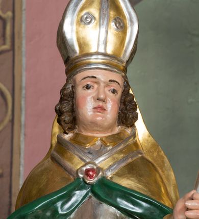 Zdjęcie nr 7: Figura przedstawiająca św. Stanisława polichromowana, niemal pełnoplastyczna, wydrążona z tyłu. Święty ukazany w pozycji stojącej, w hieratycznej pozie. Głowa przechylona lekko w lewą stronę, ręce wysunięte, skierowane w lewą stronę, w lewej dłoni święty trzyma pastorał. Twarz szeroka, schematycznie opracowana, z puklami ciemnobrązowych włosów po bokach. Święty jest ubrany w szaty biskupie: czerwoną sutannę, srebrną komżę oraz złotą kapę z zielonym podszyciem spiętą na piersi broszą, na głowie infuła.