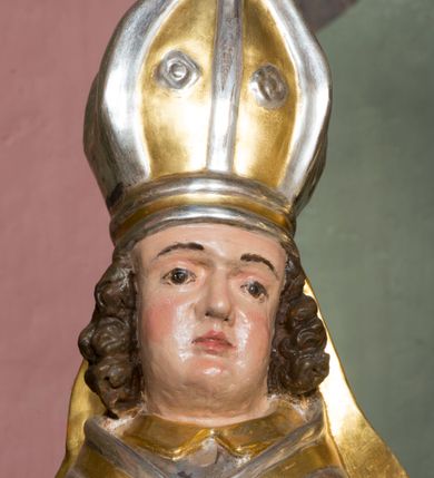 Zdjęcie nr 9: Figura przedstawiająca św. Stanisława polichromowana, niemal pełnoplastyczna, wydrążona z tyłu. Święty ukazany w pozycji stojącej, w hieratycznej pozie. Głowa przechylona lekko w lewą stronę, ręce wysunięte, skierowane w lewą stronę, w lewej dłoni święty trzyma pastorał. Twarz szeroka, schematycznie opracowana, z puklami ciemnobrązowych włosów po bokach. Święty jest ubrany w szaty biskupie: czerwoną sutannę, srebrną komżę oraz złotą kapę z zielonym podszyciem spiętą na piersi broszą, na głowie infuła.