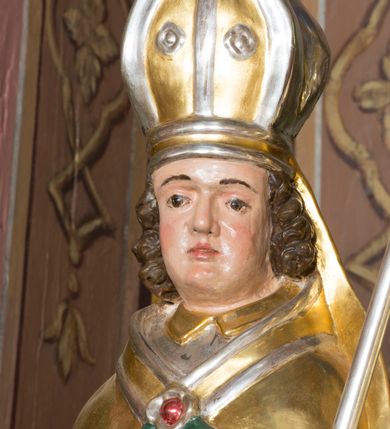 Zdjęcie nr 8: Figura przedstawiająca św. Stanisława polichromowana, niemal pełnoplastyczna, wydrążona z tyłu. Święty ukazany w pozycji stojącej, w hieratycznej pozie. Głowa przechylona lekko w lewą stronę, ręce wysunięte, skierowane w lewą stronę, w lewej dłoni święty trzyma pastorał. Twarz szeroka, schematycznie opracowana, z puklami ciemnobrązowych włosów po bokach. Święty jest ubrany w szaty biskupie: czerwoną sutannę, srebrną komżę oraz złotą kapę z zielonym podszyciem spiętą na piersi broszą, na głowie infuła.