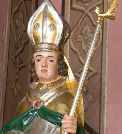 Zdjęcie nr 6: Figura przedstawiająca św. Stanisława polichromowana, niemal pełnoplastyczna, wydrążona z tyłu. Święty ukazany w pozycji stojącej, w hieratycznej pozie. Głowa przechylona lekko w lewą stronę, ręce wysunięte, skierowane w lewą stronę, w lewej dłoni święty trzyma pastorał. Twarz szeroka, schematycznie opracowana, z puklami ciemnobrązowych włosów po bokach. Święty jest ubrany w szaty biskupie: czerwoną sutannę, srebrną komżę oraz złotą kapę z zielonym podszyciem spiętą na piersi broszą, na głowie infuła.