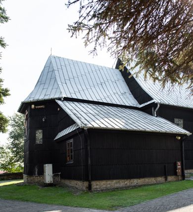 Zdjęcie nr 19: Kościół drewniany o konstrukcji zrębowej, elewacje jednolite, szalowane gontem. Wieża i kruchta zachodnia o konstrukcji słupowo-ramowej, narożne słupy nośne wzmocnione krzyżami św. Andrzeja. Więźba dachowa drewniana, krokwiowa. Budynek na kamiennym podmurowaniu. 
Kościół orientowany, jednonawowy, nawa na rzucie prostokąta zbliżonego do kwadratu, prezbiterium nieco węższe, wydłużone, zakończone trójbocznie. Do północnej ściany prezbiterium przylega zakrystia na planie prostokąta. Przy zachodniej ścianie nawy wieża na planie zbliżonym do prostokąta, o lekko ukośnej ścianie południowej. Od zachodu do wieży przylega kruchta na rzucie prostokąta. 
W przyziemiu wieży babiniec połączony z nawą wtórnym otworem w ścianie. Pomiędzy nawą a prezbiterium otwór tęczowy. Parapet chóru muzycznego w zachodniej części nawy, wsparty na czterech drewnianych kolumnach, z wejściem z nawy po drabiniastych schodach. Ściany i sklepienia pokryte polichromią z iluzjonistyczną architekturą i scenami figuralnymi. Wnętrze nakryte pułapami deskowymi w nawie i prezbiterium oraz stropem belkowym w babińcu. Otwory drzwiowe o prostokątnym wykroju zamknięte drzwiami obitymi deskami w kruchcie zachodniej, w ścianie południowej nawy oraz z prezbiterium do zakrystii. Okna w formie stojącego prostokąta. Posadzka kamienna, z płyt marmurowych. 
Elewacja frontowa jednoosiowa. Na osi wejście do kruchty zachodniej. Korpus wieży podzielony poziomo daszkami okapowymi, na froncie okno. Elewacje boczne jednolite. Korpus nawowy przepruty oknami po dwa na ścianach północnej i południowej, prezbiterium z oknem na ścianie południowej oraz dwoma okienkami na ścianach zamknięcia. Na elewacji wschodniej zawieszony krucyfiks. 
Kościół nakryty dachami dwuspadowymi, o pokryciu blaszanym, wyższym nad nawą, niższym nad prezbiterium. W połowie długości dachu, nad nawą, wbudowana wieżyczka na sygnaturkę, na ośmiobocznym, obitym blachą trzonie, z latarnią zwieńczoną niewielkim hełmem. Wieża nakryta czterospadowym daszkiem namiotowym, zwieńczona kulistą puszką i krzyżem. Kruchta nakryta osobnym daszkiem dwuspadowym, półszczytowym, zakrystia niższym dachem pulpitowym.
