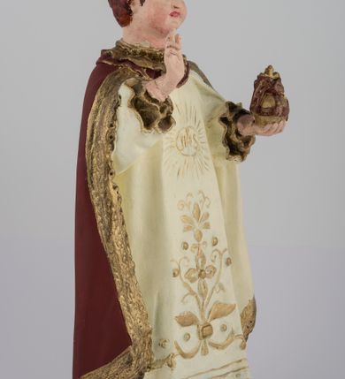 Zdjęcie nr 2: Rzeźba pełnoplastyczna, przedstawiająca stojące na profilowanej podstawie Dzieciątko Jezus. Wzrok skierowany przed siebie, nos mały, prosty, usta pełne, kości policzkowe wydatne. Włosy brązowe, na głowie bordowa, zamknięta korona ze złotymi elementami. Prawa ręka wyciągnięta w geście błogosławieństwa, w lewej królewskie jabłko. Tunika biała z długimi rękawami, zdobiona monogramem IHS z promieniami na piersi, wicią kwiatową oraz złotą lamówką. Płaszcz bordowy ze złotym wykończeniem spięty pod szyją. Szaty płasko fałdowane. Polichromia w partiach ciała naturalistyczna.