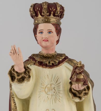 Zdjęcie nr 1: Rzeźba pełnoplastyczna, przedstawiająca stojące na profilowanej podstawie Dzieciątko Jezus. Wzrok skierowany przed siebie, nos mały, prosty, usta pełne, kości policzkowe wydatne. Włosy brązowe, na głowie bordowa, zamknięta korona ze złotymi elementami. Prawa ręka wyciągnięta w geście błogosławieństwa, w lewej królewskie jabłko. Tunika biała z długimi rękawami, zdobiona monogramem IHS z promieniami na piersi, wicią kwiatową oraz złotą lamówką. Płaszcz bordowy ze złotym wykończeniem spięty pod szyją. Szaty płasko fałdowane. Polichromia w partiach ciała naturalistyczna.