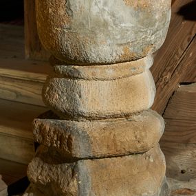 Zdjęcie nr 1: Kropielnica wmurowana w posadzkę. Trzon złożony z trzech, ustawionych na sobie fragmentów kamienia, dwa dolne są w niewielkim stopniu obrobione, górny złożony mniejszego i większego wałka, fakturowany. Czara o zarysie koła, wysoka, fakturowana. Kropielnica w kolorze piaskowca, a którego została wykonana, ubytki uzupełniono szarą zaprawą. 
