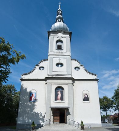 Zdjęcie nr 1: Kościół murowany, jednonawowy, na rzucie prostokąta, z późniejszymi dobudówkami. Prezbiterium usytuowane od północy, jednoprzęsłowe, węższe od korpusu, zamknięte półkolistą absydą. Do prezbiterium dobudowana zakrystia od wschodu oraz kaplica od zachodu poprzedzona niewielką kruchtą. Nawa główna trójprzęsłowa z partią wieżową niewyróżniającą się w bryle. Po bokach przęsła środkowego dostawione dwie kaplice na rzucie kwadratu: od wschodu Matki Boskiej Częstochowskiej, od zachodu św. Maksymiliana Marii Kolbego. Chór muzyczny wsparty na trzech arkadach. W prezbiterium sklepienie żaglaste, a w nawie krzyżowe, pozorne, oddzielone gurtami wychodzącymi z potężnych filarów przyściennych. Okna prostokątne, zamknięte półkoliście znajdują się w nawie, kaplicy przy prezbiterium, w wieży i w ścianach bocznych absydy. Natomiast w ścianach bocznych prezbiterium, na osi absydy i w kaplicach przy nawie okna koliste. Okna w większości wypełnione witrażami ze scenami figuralnymi, w innych ozdobne przeszklenia geometryczne. Fasada trójosiowa, na osi środkowej usytuowana wieża, delikatnie wystająca przed lico fasady, o podziałach ramowych, nakryta baniastym hełmem z dwukondygnacyjną latarnią. Osie boczne zwieńczone spływami wolutowymi. Dachy gontowe.

