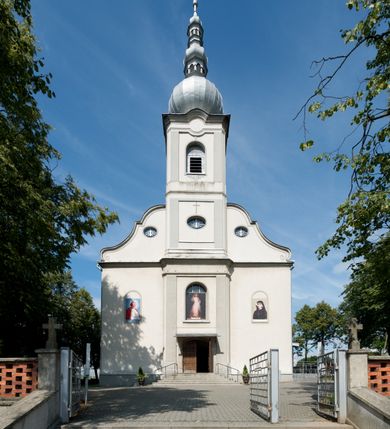 Zdjęcie nr 2: Kościół murowany, jednonawowy, na rzucie prostokąta, z późniejszymi dobudówkami. Prezbiterium usytuowane od północy, jednoprzęsłowe, węższe od korpusu, zamknięte półkolistą absydą. Do prezbiterium dobudowana zakrystia od wschodu oraz kaplica od zachodu poprzedzona niewielką kruchtą. Nawa główna trójprzęsłowa z partią wieżową niewyróżniającą się w bryle. Po bokach przęsła środkowego dostawione dwie kaplice na rzucie kwadratu: od wschodu Matki Boskiej Częstochowskiej, od zachodu św. Maksymiliana Marii Kolbego. Chór muzyczny wsparty na trzech arkadach. W prezbiterium sklepienie żaglaste, a w nawie krzyżowe, pozorne, oddzielone gurtami wychodzącymi z potężnych filarów przyściennych. Okna prostokątne, zamknięte półkoliście znajdują się w nawie, kaplicy przy prezbiterium, w wieży i w ścianach bocznych absydy. Natomiast w ścianach bocznych prezbiterium, na osi absydy i w kaplicach przy nawie okna koliste. Okna w większości wypełnione witrażami ze scenami figuralnymi, w innych ozdobne przeszklenia geometryczne. Fasada trójosiowa, na osi środkowej usytuowana wieża, delikatnie wystająca przed lico fasady, o podziałach ramowych, nakryta baniastym hełmem z dwukondygnacyjną latarnią. Osie boczne zwieńczone spływami wolutowymi. Dachy gontowe.

