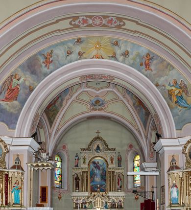 Zdjęcie nr 19: Kościół murowany, jednonawowy, na rzucie prostokąta, z późniejszymi dobudówkami. Prezbiterium usytuowane od północy, jednoprzęsłowe, węższe od korpusu, zamknięte półkolistą absydą. Do prezbiterium dobudowana zakrystia od wschodu oraz kaplica od zachodu poprzedzona niewielką kruchtą. Nawa główna trójprzęsłowa z partią wieżową niewyróżniającą się w bryle. Po bokach przęsła środkowego dostawione dwie kaplice na rzucie kwadratu: od wschodu Matki Boskiej Częstochowskiej, od zachodu św. Maksymiliana Marii Kolbego. Chór muzyczny wsparty na trzech arkadach. W prezbiterium sklepienie żaglaste, a w nawie krzyżowe, pozorne, oddzielone gurtami wychodzącymi z potężnych filarów przyściennych. Okna prostokątne, zamknięte półkoliście znajdują się w nawie, kaplicy przy prezbiterium, w wieży i w ścianach bocznych absydy. Natomiast w ścianach bocznych prezbiterium, na osi absydy i w kaplicach przy nawie okna koliste. Okna w większości wypełnione witrażami ze scenami figuralnymi, w innych ozdobne przeszklenia geometryczne. Fasada trójosiowa, na osi środkowej usytuowana wieża, delikatnie wystająca przed lico fasady, o podziałach ramowych, nakryta baniastym hełmem z dwukondygnacyjną latarnią. Osie boczne zwieńczone spływami wolutowymi. Dachy gontowe.

