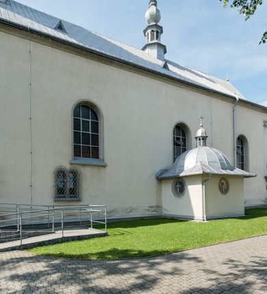 Zdjęcie nr 7: Kościół murowany, jednonawowy, na rzucie prostokąta, z późniejszymi dobudówkami. Prezbiterium usytuowane od północy, jednoprzęsłowe, węższe od korpusu, zamknięte półkolistą absydą. Do prezbiterium dobudowana zakrystia od wschodu oraz kaplica od zachodu poprzedzona niewielką kruchtą. Nawa główna trójprzęsłowa z partią wieżową niewyróżniającą się w bryle. Po bokach przęsła środkowego dostawione dwie kaplice na rzucie kwadratu: od wschodu Matki Boskiej Częstochowskiej, od zachodu św. Maksymiliana Marii Kolbego. Chór muzyczny wsparty na trzech arkadach. W prezbiterium sklepienie żaglaste, a w nawie krzyżowe, pozorne, oddzielone gurtami wychodzącymi z potężnych filarów przyściennych. Okna prostokątne, zamknięte półkoliście znajdują się w nawie, kaplicy przy prezbiterium, w wieży i w ścianach bocznych absydy. Natomiast w ścianach bocznych prezbiterium, na osi absydy i w kaplicach przy nawie okna koliste. Okna w większości wypełnione witrażami ze scenami figuralnymi, w innych ozdobne przeszklenia geometryczne. Fasada trójosiowa, na osi środkowej usytuowana wieża, delikatnie wystająca przed lico fasady, o podziałach ramowych, nakryta baniastym hełmem z dwukondygnacyjną latarnią. Osie boczne zwieńczone spływami wolutowymi. Dachy gontowe.

