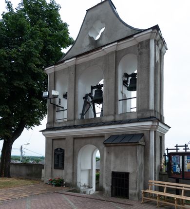 Zdjęcie nr 2: Dzwonnica parawanowa, murowana z cegły i tynkowana, trójosiowa, dwukondygnacyjna ze zwieńczeniem. Usytuowana na niewielkim wzniesieniu, w południowej części muru otaczającego kościół. Od południowego wschodu prowadzą do niej schody, otoczone murem. Dolna kondygnacja na wysokim cokole, jednoprzelotowa z usytuowanym na osi, zamkniętym półkoliście otworem przejściowym, umieszczonym w prostokątnej płycinie. Na jego osi w fasadzie południowej, muszla palmetowa. Osie skrajne z analogicznymi płycinami, w których umieszczono zamknięte półkoliście, głębokie nisze o profilowanych krawędziach. W elewacji od strony kościoła skrajne osie z prostokątnymi płycinami, po prawej, usytuowana niewielka przybudówka kryta dachem pulpitowym, z okratowanym otworem okiennym w przyziemiu. Wąskie elewacje boczne artykułowane prostokątnymi płycinami. Kondygnacja zamknięta bogato profilowanym, wydatnym gzymsem przełamującym się na bocznych krawędziach budynku. Kondygnacja druga trójprzelotowa z umieszczonymi w osiach, zamkniętymi półkoliście otworami, w miejscu których zamontowane dzwony. Elewacje południowa i północna artykułowane za pomocą pilastrów o kapitelach impostowych, łączących się z gzymsem koronującym. Zwieńczenie w kształcie łuku kotarowego o ściętym wierzchołku, z umieszczonym w jego centrum otworem o formie czwórliścia. Całość zwieńczona metalowym krzyżem. Gzymsy oddzielające kondygnacje oraz zwieńczenie kryte blachą. Tynk szary, zróżnicowany kolorystycznie bielą użytą w celu podkreślenia detalu architektonicznego.