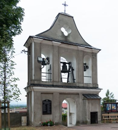 Zdjęcie nr 3: Dzwonnica parawanowa, murowana z cegły i tynkowana, trójosiowa, dwukondygnacyjna ze zwieńczeniem. Usytuowana na niewielkim wzniesieniu, w południowej części muru otaczającego kościół. Od południowego wschodu prowadzą do niej schody, otoczone murem. Dolna kondygnacja na wysokim cokole, jednoprzelotowa z usytuowanym na osi, zamkniętym półkoliście otworem przejściowym, umieszczonym w prostokątnej płycinie. Na jego osi w fasadzie południowej, muszla palmetowa. Osie skrajne z analogicznymi płycinami, w których umieszczono zamknięte półkoliście, głębokie nisze o profilowanych krawędziach. W elewacji od strony kościoła skrajne osie z prostokątnymi płycinami, po prawej, usytuowana niewielka przybudówka kryta dachem pulpitowym, z okratowanym otworem okiennym w przyziemiu. Wąskie elewacje boczne artykułowane prostokątnymi płycinami. Kondygnacja zamknięta bogato profilowanym, wydatnym gzymsem przełamującym się na bocznych krawędziach budynku. Kondygnacja druga trójprzelotowa z umieszczonymi w osiach, zamkniętymi półkoliście otworami, w miejscu których zamontowane dzwony. Elewacje południowa i północna artykułowane za pomocą pilastrów o kapitelach impostowych, łączących się z gzymsem koronującym. Zwieńczenie w kształcie łuku kotarowego o ściętym wierzchołku, z umieszczonym w jego centrum otworem o formie czwórliścia. Całość zwieńczona metalowym krzyżem. Gzymsy oddzielające kondygnacje oraz zwieńczenie kryte blachą. Tynk szary, zróżnicowany kolorystycznie bielą użytą w celu podkreślenia detalu architektonicznego.