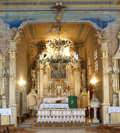 Zdjęcie nr 2: Kościół zlokalizowany na zachodnim krańcu wsi. Plac kościelny otoczony drewnianym płotem. W północno-wschodnim narożu drewniana dzwonnica.  
Kościół orientowany, drewniany, konstrukcji zrębowej, z wieżą na słup od zachodu, wejście główne od zachodu; wejścia boczne od południa przez kruchtę i od północy przez zakrystię. Prezbiterium zamknięte wielobocznie, nawa na rzucie prostokąta. Przy prezbiterium i nawie od północy zakrystia z przedsionkiem, przy nawie od południa kruchta. Wewnątrz stropy, podkreślone profilowanym gzymsem. Łuk tęczowy o wykroju prostokątnym ze ściętymi narożami, flankowany filarkami z belkowaniem; na nim późnobarokowy krucyfiks podtrzymywany przez aniołka, ponad belkowaniem aniołki dzierżące narzędzia Męki Pańskiej. Drugi łuk tęczowy usytuowany na granicy przedłużenia naw o podobnym wykroju. Chór muzyczny wsparty na dwóch żłobkowanych słupach o falistym przebiegu. Ściany nawy przeprute: od północy oknem o kształcie prostokąta, od południa trzema analogicznymi oknami i zwieńczone profilowanym gzymsem. Obramienia okienne fazowane, z nadokiennikami o wykroju w ośli grzbiet. Drzwi prowadzące do kruchty w kształcie prostokąta. Prezbiterium wzniesione o jeden stopień powyżej poziomu nawy, od południa przeprute dwoma prostokątnymi oknami. Od północy drzwi do zakrystii o kształcie stojącego prostokąta, od strony zakrystii profilowane. Zakrystia na planie prostokąta, przylegająca od północy do prezbiterium kościoła. Nawa i kruchta kryte dachem dwuspadowym, prezbiterium trójpołaciowym, opadającym ku wschodowi, zakrystia dachem pulpitowym. Przy wschodnim krańcu dachu nawy znajduje się wieżyczka na sygnaturkę. Dzwonnica drewniana, dwukondygnacyjna na planie zbliżonym do kwadratu. Nad pierwszą kondygnacją dach okapowy, a nad drugą czterospadowy, zwieńczony kulą z krzyżem. Na fasadzie kruchty wnęka z rzeźbą Matki Boskiej z Dzieciątkiem. 


