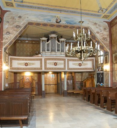Zdjęcie nr 8: Kościół zlokalizowany na zachodnim krańcu wsi. Plac kościelny otoczony drewnianym płotem. W północno-wschodnim narożu drewniana dzwonnica.  
Kościół orientowany, drewniany, konstrukcji zrębowej, z wieżą na słup od zachodu, wejście główne od zachodu; wejścia boczne od południa przez kruchtę i od północy przez zakrystię. Prezbiterium zamknięte wielobocznie, nawa na rzucie prostokąta. Przy prezbiterium i nawie od północy zakrystia z przedsionkiem, przy nawie od południa kruchta. Wewnątrz stropy, podkreślone profilowanym gzymsem. Łuk tęczowy o wykroju prostokątnym ze ściętymi narożami, flankowany filarkami z belkowaniem; na nim późnobarokowy krucyfiks podtrzymywany przez aniołka, ponad belkowaniem aniołki dzierżące narzędzia Męki Pańskiej. Drugi łuk tęczowy usytuowany na granicy przedłużenia naw o podobnym wykroju. Chór muzyczny wsparty na dwóch żłobkowanych słupach o falistym przebiegu. Ściany nawy przeprute: od północy oknem o kształcie prostokąta, od południa trzema analogicznymi oknami i zwieńczone profilowanym gzymsem. Obramienia okienne fazowane, z nadokiennikami o wykroju w ośli grzbiet. Drzwi prowadzące do kruchty w kształcie prostokąta. Prezbiterium wzniesione o jeden stopień powyżej poziomu nawy, od południa przeprute dwoma prostokątnymi oknami. Od północy drzwi do zakrystii o kształcie stojącego prostokąta, od strony zakrystii profilowane. Zakrystia na planie prostokąta, przylegająca od północy do prezbiterium kościoła. Nawa i kruchta kryte dachem dwuspadowym, prezbiterium trójpołaciowym, opadającym ku wschodowi, zakrystia dachem pulpitowym. Przy wschodnim krańcu dachu nawy znajduje się wieżyczka na sygnaturkę. Dzwonnica drewniana, dwukondygnacyjna na planie zbliżonym do kwadratu. Nad pierwszą kondygnacją dach okapowy, a nad drugą czterospadowy, zwieńczony kulą z krzyżem. Na fasadzie kruchty wnęka z rzeźbą Matki Boskiej z Dzieciątkiem. 


