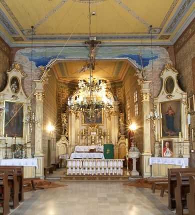 Zdjęcie nr 3: Kościół zlokalizowany na zachodnim krańcu wsi. Plac kościelny otoczony drewnianym płotem. W północno-wschodnim narożu drewniana dzwonnica.  
Kościół orientowany, drewniany, konstrukcji zrębowej, z wieżą na słup od zachodu, wejście główne od zachodu; wejścia boczne od południa przez kruchtę i od północy przez zakrystię. Prezbiterium zamknięte wielobocznie, nawa na rzucie prostokąta. Przy prezbiterium i nawie od północy zakrystia z przedsionkiem, przy nawie od południa kruchta. Wewnątrz stropy, podkreślone profilowanym gzymsem. Łuk tęczowy o wykroju prostokątnym ze ściętymi narożami, flankowany filarkami z belkowaniem; na nim późnobarokowy krucyfiks podtrzymywany przez aniołka, ponad belkowaniem aniołki dzierżące narzędzia Męki Pańskiej. Drugi łuk tęczowy usytuowany na granicy przedłużenia naw o podobnym wykroju. Chór muzyczny wsparty na dwóch żłobkowanych słupach o falistym przebiegu. Ściany nawy przeprute: od północy oknem o kształcie prostokąta, od południa trzema analogicznymi oknami i zwieńczone profilowanym gzymsem. Obramienia okienne fazowane, z nadokiennikami o wykroju w ośli grzbiet. Drzwi prowadzące do kruchty w kształcie prostokąta. Prezbiterium wzniesione o jeden stopień powyżej poziomu nawy, od południa przeprute dwoma prostokątnymi oknami. Od północy drzwi do zakrystii o kształcie stojącego prostokąta, od strony zakrystii profilowane. Zakrystia na planie prostokąta, przylegająca od północy do prezbiterium kościoła. Nawa i kruchta kryte dachem dwuspadowym, prezbiterium trójpołaciowym, opadającym ku wschodowi, zakrystia dachem pulpitowym. Przy wschodnim krańcu dachu nawy znajduje się wieżyczka na sygnaturkę. Dzwonnica drewniana, dwukondygnacyjna na planie zbliżonym do kwadratu. Nad pierwszą kondygnacją dach okapowy, a nad drugą czterospadowy, zwieńczony kulą z krzyżem. Na fasadzie kruchty wnęka z rzeźbą Matki Boskiej z Dzieciątkiem. 


