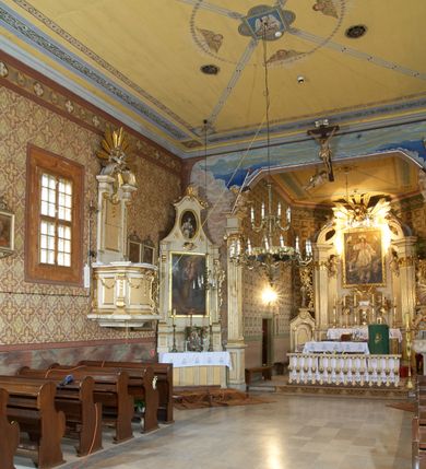 Zdjęcie nr 6: Kościół zlokalizowany na zachodnim krańcu wsi. Plac kościelny otoczony drewnianym płotem. W północno-wschodnim narożu drewniana dzwonnica.  
Kościół orientowany, drewniany, konstrukcji zrębowej, z wieżą na słup od zachodu, wejście główne od zachodu; wejścia boczne od południa przez kruchtę i od północy przez zakrystię. Prezbiterium zamknięte wielobocznie, nawa na rzucie prostokąta. Przy prezbiterium i nawie od północy zakrystia z przedsionkiem, przy nawie od południa kruchta. Wewnątrz stropy, podkreślone profilowanym gzymsem. Łuk tęczowy o wykroju prostokątnym ze ściętymi narożami, flankowany filarkami z belkowaniem; na nim późnobarokowy krucyfiks podtrzymywany przez aniołka, ponad belkowaniem aniołki dzierżące narzędzia Męki Pańskiej. Drugi łuk tęczowy usytuowany na granicy przedłużenia naw o podobnym wykroju. Chór muzyczny wsparty na dwóch żłobkowanych słupach o falistym przebiegu. Ściany nawy przeprute: od północy oknem o kształcie prostokąta, od południa trzema analogicznymi oknami i zwieńczone profilowanym gzymsem. Obramienia okienne fazowane, z nadokiennikami o wykroju w ośli grzbiet. Drzwi prowadzące do kruchty w kształcie prostokąta. Prezbiterium wzniesione o jeden stopień powyżej poziomu nawy, od południa przeprute dwoma prostokątnymi oknami. Od północy drzwi do zakrystii o kształcie stojącego prostokąta, od strony zakrystii profilowane. Zakrystia na planie prostokąta, przylegająca od północy do prezbiterium kościoła. Nawa i kruchta kryte dachem dwuspadowym, prezbiterium trójpołaciowym, opadającym ku wschodowi, zakrystia dachem pulpitowym. Przy wschodnim krańcu dachu nawy znajduje się wieżyczka na sygnaturkę. Dzwonnica drewniana, dwukondygnacyjna na planie zbliżonym do kwadratu. Nad pierwszą kondygnacją dach okapowy, a nad drugą czterospadowy, zwieńczony kulą z krzyżem. Na fasadzie kruchty wnęka z rzeźbą Matki Boskiej z Dzieciątkiem. 


