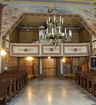 Zdjęcie nr 9: Kościół zlokalizowany na zachodnim krańcu wsi. Plac kościelny otoczony drewnianym płotem. W północno-wschodnim narożu drewniana dzwonnica.  
Kościół orientowany, drewniany, konstrukcji zrębowej, z wieżą na słup od zachodu, wejście główne od zachodu; wejścia boczne od południa przez kruchtę i od północy przez zakrystię. Prezbiterium zamknięte wielobocznie, nawa na rzucie prostokąta. Przy prezbiterium i nawie od północy zakrystia z przedsionkiem, przy nawie od południa kruchta. Wewnątrz stropy, podkreślone profilowanym gzymsem. Łuk tęczowy o wykroju prostokątnym ze ściętymi narożami, flankowany filarkami z belkowaniem; na nim późnobarokowy krucyfiks podtrzymywany przez aniołka, ponad belkowaniem aniołki dzierżące narzędzia Męki Pańskiej. Drugi łuk tęczowy usytuowany na granicy przedłużenia naw o podobnym wykroju. Chór muzyczny wsparty na dwóch żłobkowanych słupach o falistym przebiegu. Ściany nawy przeprute: od północy oknem o kształcie prostokąta, od południa trzema analogicznymi oknami i zwieńczone profilowanym gzymsem. Obramienia okienne fazowane, z nadokiennikami o wykroju w ośli grzbiet. Drzwi prowadzące do kruchty w kształcie prostokąta. Prezbiterium wzniesione o jeden stopień powyżej poziomu nawy, od południa przeprute dwoma prostokątnymi oknami. Od północy drzwi do zakrystii o kształcie stojącego prostokąta, od strony zakrystii profilowane. Zakrystia na planie prostokąta, przylegająca od północy do prezbiterium kościoła. Nawa i kruchta kryte dachem dwuspadowym, prezbiterium trójpołaciowym, opadającym ku wschodowi, zakrystia dachem pulpitowym. Przy wschodnim krańcu dachu nawy znajduje się wieżyczka na sygnaturkę. Dzwonnica drewniana, dwukondygnacyjna na planie zbliżonym do kwadratu. Nad pierwszą kondygnacją dach okapowy, a nad drugą czterospadowy, zwieńczony kulą z krzyżem. Na fasadzie kruchty wnęka z rzeźbą Matki Boskiej z Dzieciątkiem. 


