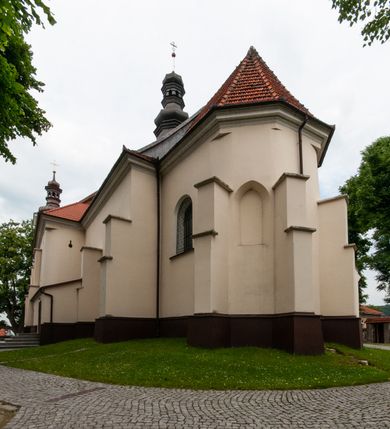 Zdjęcie nr 33: Kościół orientowany, murowany, jednonawowy z prezbiterium zakończonym wielobocznie. Wschodnia część węższa, prostokątna, zachodnia szersza, trójnawowa, halowa. Do prezbiterium od północy przylega zakrystia, do wschodniej części korpusu przybudówka, od zachodu kruchta. Ściany nawy dzielą pilastry. Prezbiterium sklepione krzyżowo, absyda konchowo z lunetami, nawy kolebkowo z lunetami. Od zachodu chór wsparty na dwóch czworobocznych słupach. Jego parapet jest wybrzuszony w części środkowej, ażurowy, zakończony u dołu listwą wycinaną w formie stylizowanych listków. W prezbiterium jedno okno w kształcie stojącego prostokąta zamknięte łukiem pełnym i jedno ślepe, zamknięte łukiem ostrym. Od strony  północnej do zakrystii dwa okna zamknięte łukiem odcinkowym i cztery prostokątne, dwa okna zamknięte łukiem pełnym do zachodniej części nawy; od strony południowej pięć okien zamkniętych łukiem pełnym. Główne wejście od zachodu, drugie od południa, kolejne do  zakrystii, portal z prezbiterium do zakrystii, z czarnego marmuru. Fasada dwuwieżowa, z kruchtą, do której prowadzi ciąg schodów. Kruchta prostopadłościenna, z dachem dwuspadowym, z portalem w kształcie prostokąta zamkniętego łukiem pełnym na osi między oknami powtarzającymi ten kształt. Nad kondygnacją trójkątny przyczółek z krzyżem łacińskim. Fasada dwukondygnacyjna, z wieżami wtopionymi w lico. Pierwsza kondygnacja gładka, flankowana szkarpami, zamknięta gzymsem, druga również niemal gładka, z zegarem na środku i małymi oknami w partii wież. Na osi trójkątny przyczółek. Elewacje obiega cokół, wspierają szkarpy – najgęściej ustawione wokół prezbiterium. Dachy naw dwuspadowe, nad prezbiterium dach zakończony wielobocznie, nad zakrystią pulpitowy; na kalenicy wieżyczka na sygnaturkę, wieże zakończone rozbudowanymi hełmami. 
We wnętrzu na uwagę zasługują chrzcielnica z czarnego marmuru z XVII wieku, obraz św. Izydora w prawym, bocznym ołtarzu według tradycji pędzla Michała Stachowicza i późnobarokowe ołtarze. Prowadzący do zakrystii portal z czarnego marmuru i ołtarz boczny lewy Ukrzyżowania pochodzą, według niektórych przekazów, z krakowskiego kościoła Wszystkich Świętych. 
