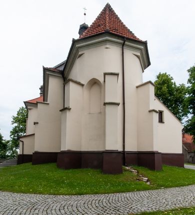 Zdjęcie nr 34: Kościół orientowany, murowany, jednonawowy z prezbiterium zakończonym wielobocznie. Wschodnia część węższa, prostokątna, zachodnia szersza, trójnawowa, halowa. Do prezbiterium od północy przylega zakrystia, do wschodniej części korpusu przybudówka, od zachodu kruchta. Ściany nawy dzielą pilastry. Prezbiterium sklepione krzyżowo, absyda konchowo z lunetami, nawy kolebkowo z lunetami. Od zachodu chór wsparty na dwóch czworobocznych słupach. Jego parapet jest wybrzuszony w części środkowej, ażurowy, zakończony u dołu listwą wycinaną w formie stylizowanych listków. W prezbiterium jedno okno w kształcie stojącego prostokąta zamknięte łukiem pełnym i jedno ślepe, zamknięte łukiem ostrym. Od strony  północnej do zakrystii dwa okna zamknięte łukiem odcinkowym i cztery prostokątne, dwa okna zamknięte łukiem pełnym do zachodniej części nawy; od strony południowej pięć okien zamkniętych łukiem pełnym. Główne wejście od zachodu, drugie od południa, kolejne do  zakrystii, portal z prezbiterium do zakrystii, z czarnego marmuru. Fasada dwuwieżowa, z kruchtą, do której prowadzi ciąg schodów. Kruchta prostopadłościenna, z dachem dwuspadowym, z portalem w kształcie prostokąta zamkniętego łukiem pełnym na osi między oknami powtarzającymi ten kształt. Nad kondygnacją trójkątny przyczółek z krzyżem łacińskim. Fasada dwukondygnacyjna, z wieżami wtopionymi w lico. Pierwsza kondygnacja gładka, flankowana szkarpami, zamknięta gzymsem, druga również niemal gładka, z zegarem na środku i małymi oknami w partii wież. Na osi trójkątny przyczółek. Elewacje obiega cokół, wspierają szkarpy – najgęściej ustawione wokół prezbiterium. Dachy naw dwuspadowe, nad prezbiterium dach zakończony wielobocznie, nad zakrystią pulpitowy; na kalenicy wieżyczka na sygnaturkę, wieże zakończone rozbudowanymi hełmami. 
We wnętrzu na uwagę zasługują chrzcielnica z czarnego marmuru z XVII wieku, obraz św. Izydora w prawym, bocznym ołtarzu według tradycji pędzla Michała Stachowicza i późnobarokowe ołtarze. Prowadzący do zakrystii portal z czarnego marmuru i ołtarz boczny lewy Ukrzyżowania pochodzą, według niektórych przekazów, z krakowskiego kościoła Wszystkich Świętych. 
