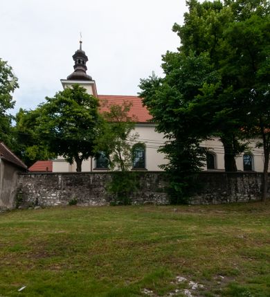 Zdjęcie nr 15: Kościół orientowany, murowany, jednonawowy z prezbiterium zakończonym wielobocznie. Wschodnia część węższa, prostokątna, zachodnia szersza, trójnawowa, halowa. Do prezbiterium od północy przylega zakrystia, do wschodniej części korpusu przybudówka, od zachodu kruchta. Ściany nawy dzielą pilastry. Prezbiterium sklepione krzyżowo, absyda konchowo z lunetami, nawy kolebkowo z lunetami. Od zachodu chór wsparty na dwóch czworobocznych słupach. Jego parapet jest wybrzuszony w części środkowej, ażurowy, zakończony u dołu listwą wycinaną w formie stylizowanych listków. W prezbiterium jedno okno w kształcie stojącego prostokąta zamknięte łukiem pełnym i jedno ślepe, zamknięte łukiem ostrym. Od strony  północnej do zakrystii dwa okna zamknięte łukiem odcinkowym i cztery prostokątne, dwa okna zamknięte łukiem pełnym do zachodniej części nawy; od strony południowej pięć okien zamkniętych łukiem pełnym. Główne wejście od zachodu, drugie od południa, kolejne do  zakrystii, portal z prezbiterium do zakrystii, z czarnego marmuru. Fasada dwuwieżowa, z kruchtą, do której prowadzi ciąg schodów. Kruchta prostopadłościenna, z dachem dwuspadowym, z portalem w kształcie prostokąta zamkniętego łukiem pełnym na osi między oknami powtarzającymi ten kształt. Nad kondygnacją trójkątny przyczółek z krzyżem łacińskim. Fasada dwukondygnacyjna, z wieżami wtopionymi w lico. Pierwsza kondygnacja gładka, flankowana szkarpami, zamknięta gzymsem, druga również niemal gładka, z zegarem na środku i małymi oknami w partii wież. Na osi trójkątny przyczółek. Elewacje obiega cokół, wspierają szkarpy – najgęściej ustawione wokół prezbiterium. Dachy naw dwuspadowe, nad prezbiterium dach zakończony wielobocznie, nad zakrystią pulpitowy; na kalenicy wieżyczka na sygnaturkę, wieże zakończone rozbudowanymi hełmami. 
We wnętrzu na uwagę zasługują chrzcielnica z czarnego marmuru z XVII wieku, obraz św. Izydora w prawym, bocznym ołtarzu według tradycji pędzla Michała Stachowicza i późnobarokowe ołtarze. Prowadzący do zakrystii portal z czarnego marmuru i ołtarz boczny lewy Ukrzyżowania pochodzą, według niektórych przekazów, z krakowskiego kościoła Wszystkich Świętych. 
