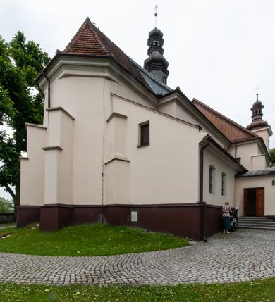 Zdjęcie nr 31: Kościół orientowany, murowany, jednonawowy z prezbiterium zakończonym wielobocznie. Wschodnia część węższa, prostokątna, zachodnia szersza, trójnawowa, halowa. Do prezbiterium od północy przylega zakrystia, do wschodniej części korpusu przybudówka, od zachodu kruchta. Ściany nawy dzielą pilastry. Prezbiterium sklepione krzyżowo, absyda konchowo z lunetami, nawy kolebkowo z lunetami. Od zachodu chór wsparty na dwóch czworobocznych słupach. Jego parapet jest wybrzuszony w części środkowej, ażurowy, zakończony u dołu listwą wycinaną w formie stylizowanych listków. W prezbiterium jedno okno w kształcie stojącego prostokąta zamknięte łukiem pełnym i jedno ślepe, zamknięte łukiem ostrym. Od strony  północnej do zakrystii dwa okna zamknięte łukiem odcinkowym i cztery prostokątne, dwa okna zamknięte łukiem pełnym do zachodniej części nawy; od strony południowej pięć okien zamkniętych łukiem pełnym. Główne wejście od zachodu, drugie od południa, kolejne do  zakrystii, portal z prezbiterium do zakrystii, z czarnego marmuru. Fasada dwuwieżowa, z kruchtą, do której prowadzi ciąg schodów. Kruchta prostopadłościenna, z dachem dwuspadowym, z portalem w kształcie prostokąta zamkniętego łukiem pełnym na osi między oknami powtarzającymi ten kształt. Nad kondygnacją trójkątny przyczółek z krzyżem łacińskim. Fasada dwukondygnacyjna, z wieżami wtopionymi w lico. Pierwsza kondygnacja gładka, flankowana szkarpami, zamknięta gzymsem, druga również niemal gładka, z zegarem na środku i małymi oknami w partii wież. Na osi trójkątny przyczółek. Elewacje obiega cokół, wspierają szkarpy – najgęściej ustawione wokół prezbiterium. Dachy naw dwuspadowe, nad prezbiterium dach zakończony wielobocznie, nad zakrystią pulpitowy; na kalenicy wieżyczka na sygnaturkę, wieże zakończone rozbudowanymi hełmami. 
We wnętrzu na uwagę zasługują chrzcielnica z czarnego marmuru z XVII wieku, obraz św. Izydora w prawym, bocznym ołtarzu według tradycji pędzla Michała Stachowicza i późnobarokowe ołtarze. Prowadzący do zakrystii portal z czarnego marmuru i ołtarz boczny lewy Ukrzyżowania pochodzą, według niektórych przekazów, z krakowskiego kościoła Wszystkich Świętych. 
