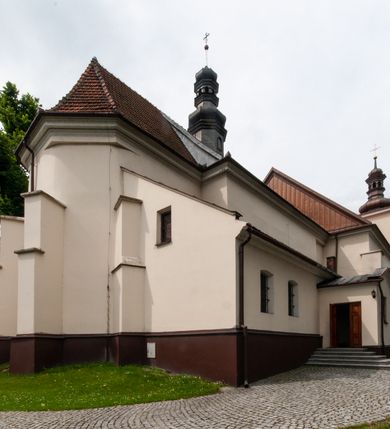 Zdjęcie nr 7: Kościół orientowany, murowany, jednonawowy z prezbiterium zakończonym wielobocznie. Wschodnia część węższa, prostokątna, zachodnia szersza, trójnawowa, halowa. Do prezbiterium od północy przylega zakrystia, do wschodniej części korpusu przybudówka, od zachodu kruchta. Ściany nawy dzielą pilastry. Prezbiterium sklepione krzyżowo, absyda konchowo z lunetami, nawy kolebkowo z lunetami. Od zachodu chór wsparty na dwóch czworobocznych słupach. Jego parapet jest wybrzuszony w części środkowej, ażurowy, zakończony u dołu listwą wycinaną w formie stylizowanych listków. W prezbiterium jedno okno w kształcie stojącego prostokąta zamknięte łukiem pełnym i jedno ślepe, zamknięte łukiem ostrym. Od strony  północnej do zakrystii dwa okna zamknięte łukiem odcinkowym i cztery prostokątne, dwa okna zamknięte łukiem pełnym do zachodniej części nawy; od strony południowej pięć okien zamkniętych łukiem pełnym. Główne wejście od zachodu, drugie od południa, kolejne do  zakrystii, portal z prezbiterium do zakrystii, z czarnego marmuru. Fasada dwuwieżowa, z kruchtą, do której prowadzi ciąg schodów. Kruchta prostopadłościenna, z dachem dwuspadowym, z portalem w kształcie prostokąta zamkniętego łukiem pełnym na osi między oknami powtarzającymi ten kształt. Nad kondygnacją trójkątny przyczółek z krzyżem łacińskim. Fasada dwukondygnacyjna, z wieżami wtopionymi w lico. Pierwsza kondygnacja gładka, flankowana szkarpami, zamknięta gzymsem, druga również niemal gładka, z zegarem na środku i małymi oknami w partii wież. Na osi trójkątny przyczółek. Elewacje obiega cokół, wspierają szkarpy – najgęściej ustawione wokół prezbiterium. Dachy naw dwuspadowe, nad prezbiterium dach zakończony wielobocznie, nad zakrystią pulpitowy; na kalenicy wieżyczka na sygnaturkę, wieże zakończone rozbudowanymi hełmami. 
We wnętrzu na uwagę zasługują chrzcielnica z czarnego marmuru z XVII wieku, obraz św. Izydora w prawym, bocznym ołtarzu według tradycji pędzla Michała Stachowicza i późnobarokowe ołtarze. Prowadzący do zakrystii portal z czarnego marmuru i ołtarz boczny lewy Ukrzyżowania pochodzą, według niektórych przekazów, z krakowskiego kościoła Wszystkich Świętych. 
