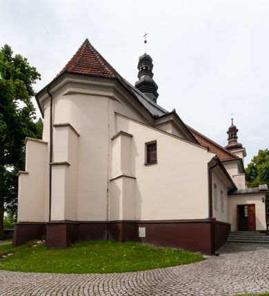 Zdjęcie nr 4: Kościół orientowany, murowany, jednonawowy z prezbiterium zakończonym wielobocznie. Wschodnia część węższa, prostokątna, zachodnia szersza, trójnawowa, halowa. Do prezbiterium od północy przylega zakrystia, do wschodniej części korpusu przybudówka, od zachodu kruchta. Ściany nawy dzielą pilastry. Prezbiterium sklepione krzyżowo, absyda konchowo z lunetami, nawy kolebkowo z lunetami. Od zachodu chór wsparty na dwóch czworobocznych słupach. Jego parapet jest wybrzuszony w części środkowej, ażurowy, zakończony u dołu listwą wycinaną w formie stylizowanych listków. W prezbiterium jedno okno w kształcie stojącego prostokąta zamknięte łukiem pełnym i jedno ślepe, zamknięte łukiem ostrym. Od strony  północnej do zakrystii dwa okna zamknięte łukiem odcinkowym i cztery prostokątne, dwa okna zamknięte łukiem pełnym do zachodniej części nawy; od strony południowej pięć okien zamkniętych łukiem pełnym. Główne wejście od zachodu, drugie od południa, kolejne do  zakrystii, portal z prezbiterium do zakrystii, z czarnego marmuru. Fasada dwuwieżowa, z kruchtą, do której prowadzi ciąg schodów. Kruchta prostopadłościenna, z dachem dwuspadowym, z portalem w kształcie prostokąta zamkniętego łukiem pełnym na osi między oknami powtarzającymi ten kształt. Nad kondygnacją trójkątny przyczółek z krzyżem łacińskim. Fasada dwukondygnacyjna, z wieżami wtopionymi w lico. Pierwsza kondygnacja gładka, flankowana szkarpami, zamknięta gzymsem, druga również niemal gładka, z zegarem na środku i małymi oknami w partii wież. Na osi trójkątny przyczółek. Elewacje obiega cokół, wspierają szkarpy – najgęściej ustawione wokół prezbiterium. Dachy naw dwuspadowe, nad prezbiterium dach zakończony wielobocznie, nad zakrystią pulpitowy; na kalenicy wieżyczka na sygnaturkę, wieże zakończone rozbudowanymi hełmami. 
We wnętrzu na uwagę zasługują chrzcielnica z czarnego marmuru z XVII wieku, obraz św. Izydora w prawym, bocznym ołtarzu według tradycji pędzla Michała Stachowicza i późnobarokowe ołtarze. Prowadzący do zakrystii portal z czarnego marmuru i ołtarz boczny lewy Ukrzyżowania pochodzą, według niektórych przekazów, z krakowskiego kościoła Wszystkich Świętych. 
