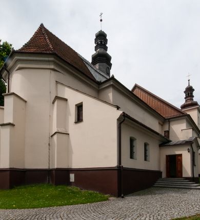 Zdjęcie nr 6: Kościół orientowany, murowany, jednonawowy z prezbiterium zakończonym wielobocznie. Wschodnia część węższa, prostokątna, zachodnia szersza, trójnawowa, halowa. Do prezbiterium od północy przylega zakrystia, do wschodniej części korpusu przybudówka, od zachodu kruchta. Ściany nawy dzielą pilastry. Prezbiterium sklepione krzyżowo, absyda konchowo z lunetami, nawy kolebkowo z lunetami. Od zachodu chór wsparty na dwóch czworobocznych słupach. Jego parapet jest wybrzuszony w części środkowej, ażurowy, zakończony u dołu listwą wycinaną w formie stylizowanych listków. W prezbiterium jedno okno w kształcie stojącego prostokąta zamknięte łukiem pełnym i jedno ślepe, zamknięte łukiem ostrym. Od strony  północnej do zakrystii dwa okna zamknięte łukiem odcinkowym i cztery prostokątne, dwa okna zamknięte łukiem pełnym do zachodniej części nawy; od strony południowej pięć okien zamkniętych łukiem pełnym. Główne wejście od zachodu, drugie od południa, kolejne do  zakrystii, portal z prezbiterium do zakrystii, z czarnego marmuru. Fasada dwuwieżowa, z kruchtą, do której prowadzi ciąg schodów. Kruchta prostopadłościenna, z dachem dwuspadowym, z portalem w kształcie prostokąta zamkniętego łukiem pełnym na osi między oknami powtarzającymi ten kształt. Nad kondygnacją trójkątny przyczółek z krzyżem łacińskim. Fasada dwukondygnacyjna, z wieżami wtopionymi w lico. Pierwsza kondygnacja gładka, flankowana szkarpami, zamknięta gzymsem, druga również niemal gładka, z zegarem na środku i małymi oknami w partii wież. Na osi trójkątny przyczółek. Elewacje obiega cokół, wspierają szkarpy – najgęściej ustawione wokół prezbiterium. Dachy naw dwuspadowe, nad prezbiterium dach zakończony wielobocznie, nad zakrystią pulpitowy; na kalenicy wieżyczka na sygnaturkę, wieże zakończone rozbudowanymi hełmami. 
We wnętrzu na uwagę zasługują chrzcielnica z czarnego marmuru z XVII wieku, obraz św. Izydora w prawym, bocznym ołtarzu według tradycji pędzla Michała Stachowicza i późnobarokowe ołtarze. Prowadzący do zakrystii portal z czarnego marmuru i ołtarz boczny lewy Ukrzyżowania pochodzą, według niektórych przekazów, z krakowskiego kościoła Wszystkich Świętych. 
