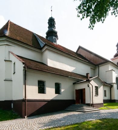 Zdjęcie nr 5: Kościół orientowany, murowany, jednonawowy z prezbiterium zakończonym wielobocznie. Wschodnia część węższa, prostokątna, zachodnia szersza, trójnawowa, halowa. Do prezbiterium od północy przylega zakrystia, do wschodniej części korpusu przybudówka, od zachodu kruchta. Ściany nawy dzielą pilastry. Prezbiterium sklepione krzyżowo, absyda konchowo z lunetami, nawy kolebkowo z lunetami. Od zachodu chór wsparty na dwóch czworobocznych słupach. Jego parapet jest wybrzuszony w części środkowej, ażurowy, zakończony u dołu listwą wycinaną w formie stylizowanych listków. W prezbiterium jedno okno w kształcie stojącego prostokąta zamknięte łukiem pełnym i jedno ślepe, zamknięte łukiem ostrym. Od strony  północnej do zakrystii dwa okna zamknięte łukiem odcinkowym i cztery prostokątne, dwa okna zamknięte łukiem pełnym do zachodniej części nawy; od strony południowej pięć okien zamkniętych łukiem pełnym. Główne wejście od zachodu, drugie od południa, kolejne do  zakrystii, portal z prezbiterium do zakrystii, z czarnego marmuru. Fasada dwuwieżowa, z kruchtą, do której prowadzi ciąg schodów. Kruchta prostopadłościenna, z dachem dwuspadowym, z portalem w kształcie prostokąta zamkniętego łukiem pełnym na osi między oknami powtarzającymi ten kształt. Nad kondygnacją trójkątny przyczółek z krzyżem łacińskim. Fasada dwukondygnacyjna, z wieżami wtopionymi w lico. Pierwsza kondygnacja gładka, flankowana szkarpami, zamknięta gzymsem, druga również niemal gładka, z zegarem na środku i małymi oknami w partii wież. Na osi trójkątny przyczółek. Elewacje obiega cokół, wspierają szkarpy – najgęściej ustawione wokół prezbiterium. Dachy naw dwuspadowe, nad prezbiterium dach zakończony wielobocznie, nad zakrystią pulpitowy; na kalenicy wieżyczka na sygnaturkę, wieże zakończone rozbudowanymi hełmami. 
We wnętrzu na uwagę zasługują chrzcielnica z czarnego marmuru z XVII wieku, obraz św. Izydora w prawym, bocznym ołtarzu według tradycji pędzla Michała Stachowicza i późnobarokowe ołtarze. Prowadzący do zakrystii portal z czarnego marmuru i ołtarz boczny lewy Ukrzyżowania pochodzą, według niektórych przekazów, z krakowskiego kościoła Wszystkich Świętych. 
