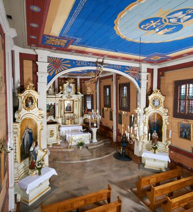 Zdjęcie nr 28: Kościół usytuowany jest w centrum wsi na niewielkim wzniesieniu, orientowany, drewniany, o konstrukcji zrębowej, oszalowany. Jego bryłę tworzy trójbocznie zamknięte prezbiterium, jednonawowy korpus, zakrystia skomunikowana ze skarbcem oraz kruchty od południa i zachodu. Całość pokrywają oddzielne dla nawy i prezbiterium dachy, nad nawą znajduje się wieżyczka na sygnaturkę. Wnętrza niższego, trójbocznie zamkniętego prezbiterium i szerszej, prawie kwadratowej nawy nakryte są stropem. Ściany kościoła zostały podzielone na przęsła kanelowanymi pilastrami ustawionymi na cokołach i zwieńczone profilowanym gzymsem, wyłamującym się na kapitelach. W prezbiterium znajdują się dwa okna od południa i dwa w bocznych ścianach wschodniego zamknięcia; korpus jest oświetlony od południa dwoma oknami. Wszystkie okna zamknięte są łukiem odcinkowym i umieszczone w prostokątnych obramieniach. Przestrzeń między prezbiterium a nawą jest nieznacznie zawężona wydatnymi pilastrami ustawionymi pod arkadą tęczową o łuku koszowym. W środkowym przęśle prezbiterium znajduje się wejście do północnej zakrystii, z profilowanym portalem zamkniętym łukiem trójlistnie ściętym. Na osi ściany południowej umieszczono prostokątny portal, ozdobiony od strony kruchty profilowaniem, a w nadprożu zdwojonym łukiem w  „ośli grzbiet”. Pod nim ornament arkadkowy i wyryta data „1547”. Główne wejście w ścianie zachodniej obejmuje profilowany od strony kruchty portal, który podobnie jak poprzedni zamknięty jest zdwojonym łukiem w „ośli grzbiet”. Chór muzyczny opiera się na dwóch kwadratowych słupach ustawionych diagonalnie (górą dekoracyjnie wycinanych), z parapetem o linii wklęsło-wypukłej. W południowo-zachodnim narożu znajdują się schody na chór osłonięte deskami. Zakrystia jest założona na rzucie prostokąta, przekryta pułapem, z lisicami na ścianie południowej i zachodniej. W osi ściany wschodniej zakrystii umieszczono okno zamknięte łukiem odcinkowym, a w narożu południowo-zachodnim schody na ambonę. W zachodniej ścianie zakrystii znajduje się prostokątne przejście do wąskiego przedsionka i dalej do dawnego skarbca przylegającego do korpusu. Kruchty są założone na rzucie prostokąta, wzdłużnie przylegającego do korpusu i przykryte stropem. Naprzeciw fasady ustawiono wolnostojącą, murowaną dzwonnicę.