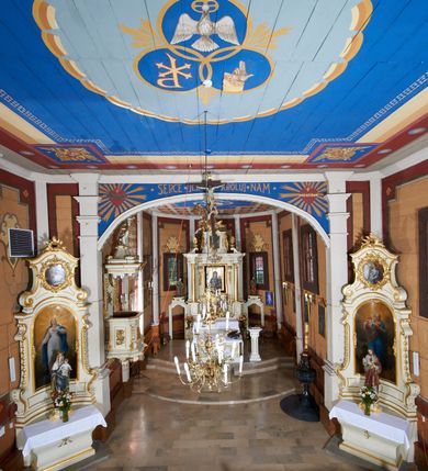 Zdjęcie nr 18: Kościół usytuowany jest w centrum wsi na niewielkim wzniesieniu, orientowany, drewniany, o konstrukcji zrębowej, oszalowany. Jego bryłę tworzy trójbocznie zamknięte prezbiterium, jednonawowy korpus, zakrystia skomunikowana ze skarbcem oraz kruchty od południa i zachodu. Całość pokrywają oddzielne dla nawy i prezbiterium dachy, nad nawą znajduje się wieżyczka na sygnaturkę. Wnętrza niższego, trójbocznie zamkniętego prezbiterium i szerszej, prawie kwadratowej nawy nakryte są stropem. Ściany kościoła zostały podzielone na przęsła kanelowanymi pilastrami ustawionymi na cokołach i zwieńczone profilowanym gzymsem, wyłamującym się na kapitelach. W prezbiterium znajdują się dwa okna od południa i dwa w bocznych ścianach wschodniego zamknięcia; korpus jest oświetlony od południa dwoma oknami. Wszystkie okna zamknięte są łukiem odcinkowym i umieszczone w prostokątnych obramieniach. Przestrzeń między prezbiterium a nawą jest nieznacznie zawężona wydatnymi pilastrami ustawionymi pod arkadą tęczową o łuku koszowym. W środkowym przęśle prezbiterium znajduje się wejście do północnej zakrystii, z profilowanym portalem zamkniętym łukiem trójlistnie ściętym. Na osi ściany południowej umieszczono prostokątny portal, ozdobiony od strony kruchty profilowaniem, a w nadprożu zdwojonym łukiem w  „ośli grzbiet”. Pod nim ornament arkadkowy i wyryta data „1547”. Główne wejście w ścianie zachodniej obejmuje profilowany od strony kruchty portal, który podobnie jak poprzedni zamknięty jest zdwojonym łukiem w „ośli grzbiet”. Chór muzyczny opiera się na dwóch kwadratowych słupach ustawionych diagonalnie (górą dekoracyjnie wycinanych), z parapetem o linii wklęsło-wypukłej. W południowo-zachodnim narożu znajdują się schody na chór osłonięte deskami. Zakrystia jest założona na rzucie prostokąta, przekryta pułapem, z lisicami na ścianie południowej i zachodniej. W osi ściany wschodniej zakrystii umieszczono okno zamknięte łukiem odcinkowym, a w narożu południowo-zachodnim schody na ambonę. W zachodniej ścianie zakrystii znajduje się prostokątne przejście do wąskiego przedsionka i dalej do dawnego skarbca przylegającego do korpusu. Kruchty są założone na rzucie prostokąta, wzdłużnie przylegającego do korpusu i przykryte stropem. Naprzeciw fasady ustawiono wolnostojącą, murowaną dzwonnicę.